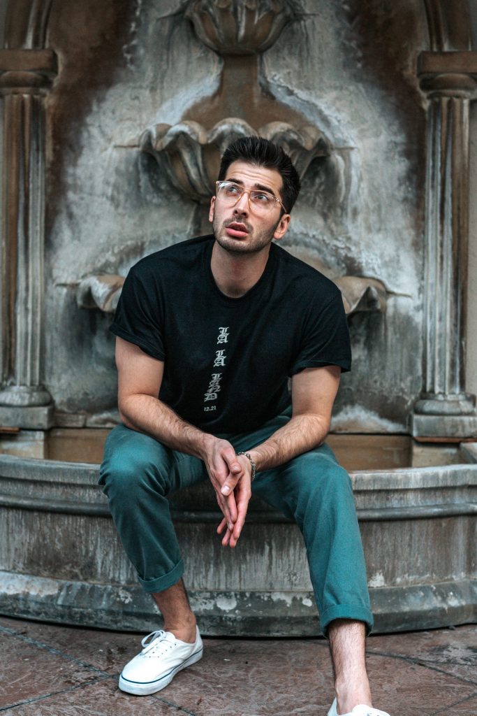 homme brun à lunettes portant un chino coloré un t shirt noir et des baskets blanches en toile