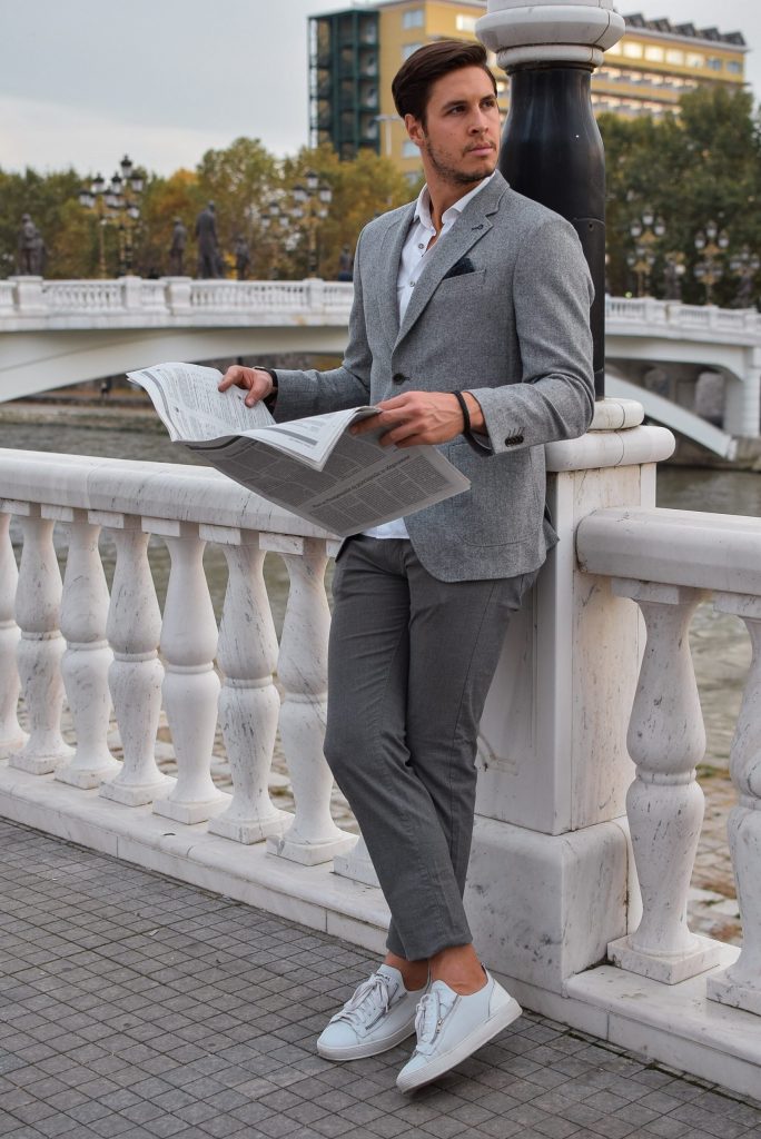 homme lisant un journal debout sur un pont portant un chino gris une veste blazer grise et des baskets blanches